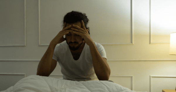 A man with long dark hair tied back sits on the edge of a bed in a dimly lit room, holding his head in his hands with a stressed expression.