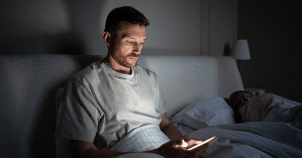 A man sitting up in bed in the dark, looking down at his glowing smartphone screen while a person slumbers beside him.
