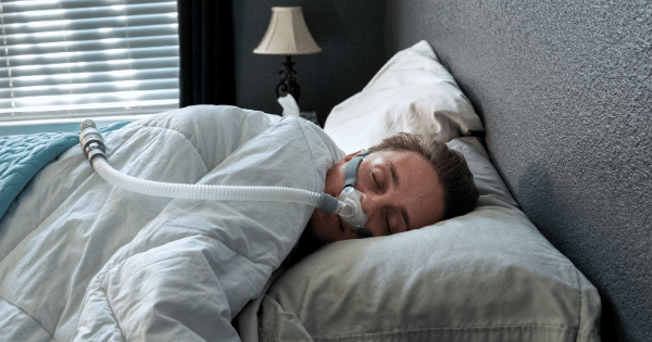 A person lying in bed on their side wearing a CPAP nasal mask attached to a white breathing hose, fast asleep under a white comforter.
