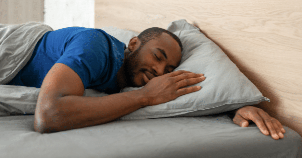 A man lying on his side on a grey bed sheet with eyes closed, resting his head on his hand over a pillow.