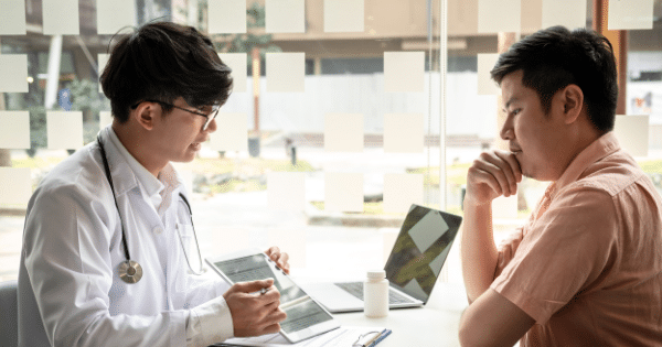 A male doctor in a white coat showing a tablet screen to a patient sitting across a table in a bright clinic office.
