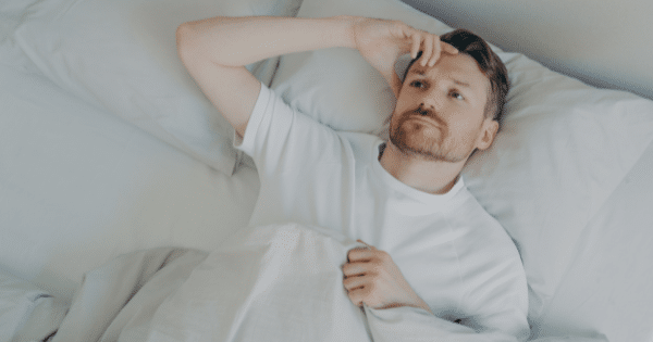 A high-angle shot of a man lying in a bed with white linens, holding one hand to his forehead with an anxious, restless expression.