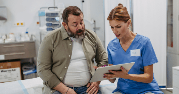 A female healthcare worker in blue scrubs holding a tablet and discussing results with a middle-aged male patient sitting on an exam table.