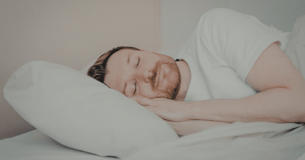 A man with a short brown beard sleeping soundly on his side with his hands resting under a plush white pillow.
