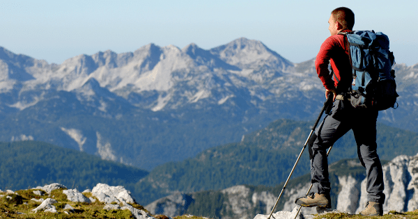 A hiker with a backpack and trekking poles standing on a rocky mountain summit, looking out toward a expansive range of distant peaks.