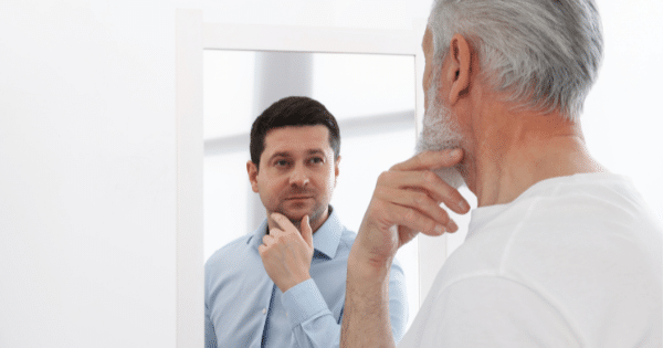 A white-haired older man looking into a mirror, touching his chin as his reflection shows a younger version of himself in a blue button-down shirt.