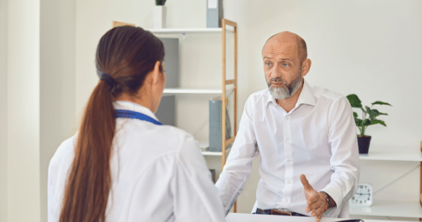 A bald man with a grey beard sitting across from a female doctor in a clinical office, gesturing with his hand as he speaks during a consultation.