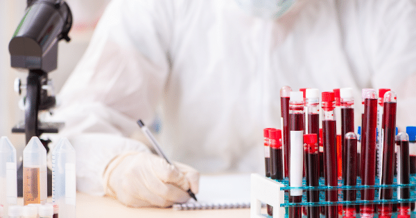A laboratory technician in protective gear writes on a notepad behind a rack of blood collection tubes, with a microscope nearby.