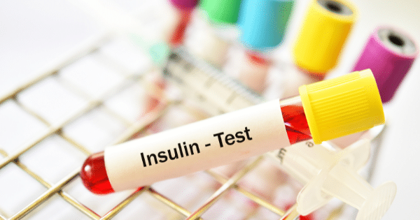 A close-up shot of a blood sample vial labeled "Insulin - Test" resting on a metal rack alongside a syringe and other colorful test tubes.