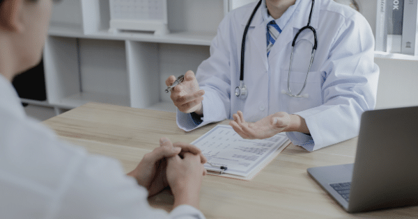 A doctor sitting at a desk with a stethoscope around their neck gestures with open hands while speaking with a patient across a table.