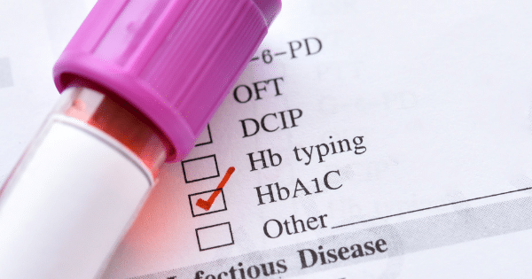 A close-up of a medical lab request form with a red checkmark next to "HbA1C" beside a purple-capped blood test tube.