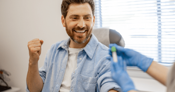 A smiling man with a beard sits in a clinic chair making a celebratory fist pump while a healthcare worker in blue gloves holds a blood sample vial.
