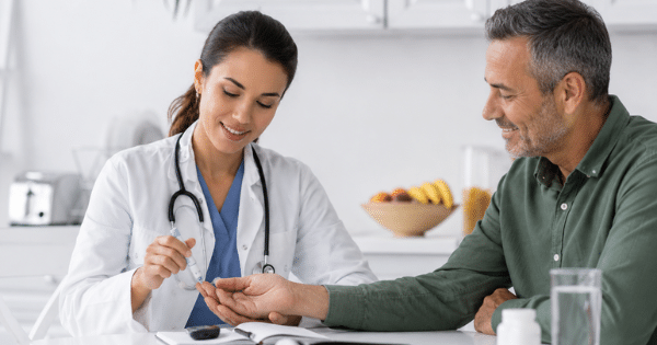 A female doctor in a white coat pricks the finger of a smiling middle-aged man to perform a blood sugar test at a table.