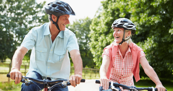 An older man and woman wearing black bicycle helmets smile at each other while riding bikes along a sunlit outdoor path.