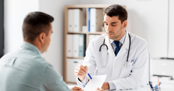 A male doctor wearing a white coat, tie, and stethoscope sits at a desk across from a patient, using a pen to point at a medical document.