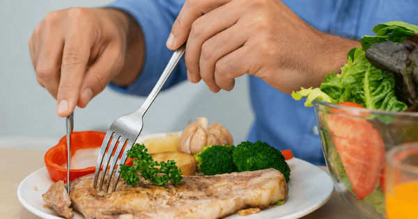 A close-up of a person using a knife and fork to cut into a cooked pork chop served with broccoli, potatoes, and a side salad.