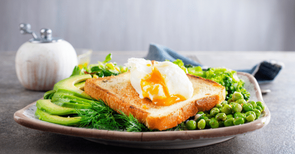 A plate featuring a slice of toasted bread topped with a runny poached egg, served with sliced avocado, green peas, and fresh greens.