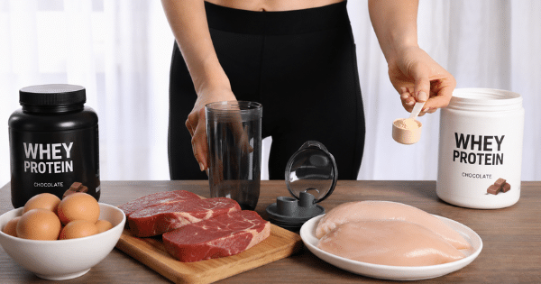 A person in black workout wear holds a scoop of powder over a shaker cup next to tubs of whey protein, raw steaks, chicken breasts, and eggs.