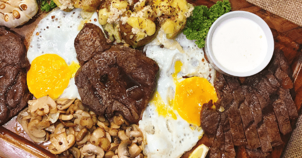 A platter filled with cooked steaks, fried sunny-side-up eggs, sautéed mushrooms, baked potatoes, and a side of white dipping sauce.