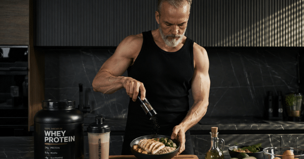 A fit, muscular older man with a grey beard wearing a black tank top grinds pepper over a healthy meal bowl in a modern kitchen, next to a whey protein tub and shaker.