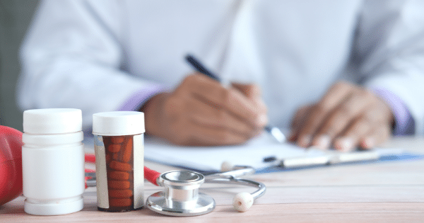 A close-up view of a doctor’s desk featuring pill bottles and a stethoscope in the foreground, with the doctor in a white coat blurred in the background writing on a document.