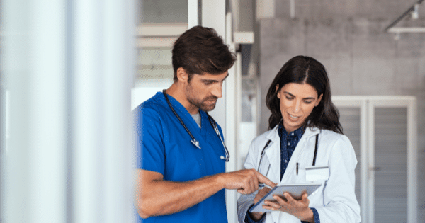 A male medical practitioner in blue scrubs and a female doctor in a white lab coat reviewing information together on a digital tablet in a hospital hallway.