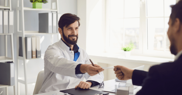 A smiling male doctor with a dark beard sitting at his office desk and handing a piece of paper or prescription to a patient.