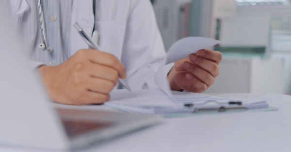 A close-up shot of a healthcare provider's hands writing with a pen on a clipboard form next to a laptop on a white desk.