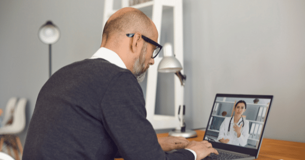 An older man with glasses viewed from behind as he participates in a virtual telehealth consultation on his laptop with a female doctor displayed on screen.