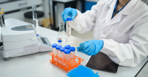 A laboratory technician wearing blue nitrile gloves and a white lab coat handling test tubes filled with clear liquid over a rack next to a digital scale.