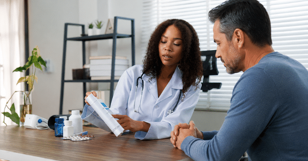 A female doctor with curly dark hair explaining a medication container to a male patient seated across her desk, with various pill bottles laid out between them.