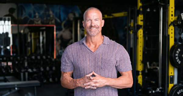 A fit, middle-aged man with short hair smiling with clasped hands while standing inside a fitness gym with weight training equipment in the background.