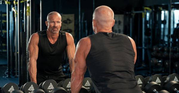 A athletic man in a black tank top leaning over a rack of dumbbells in a gym while looking intently at his reflection in a mirror.
