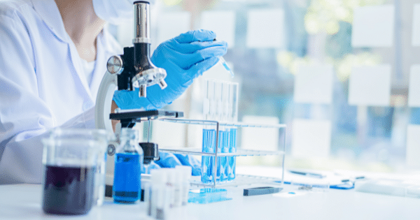 A scientist in a white lab coat and blue gloves using a pipette to add liquid to test tubes next to a compound microscope in a laboratory setting.