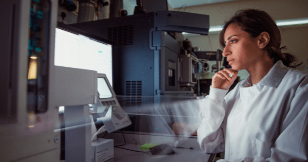 A female researcher in a white lab coat sitting thoughtfully in front of complex laboratory instruments and computer displays.
