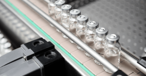 A row of small clear glass pharmaceutical vials with silver caps moving along an automated conveyor production line.