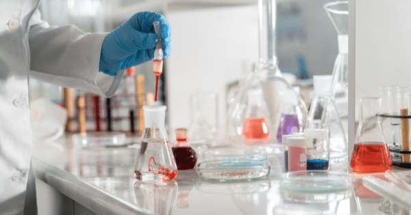 A laboratory worker wearing blue nitrile gloves uses a pipette to drop red liquid into an Erlenmeyer flask filled with clear fluid, surrounded by glassware and Petri dishes.