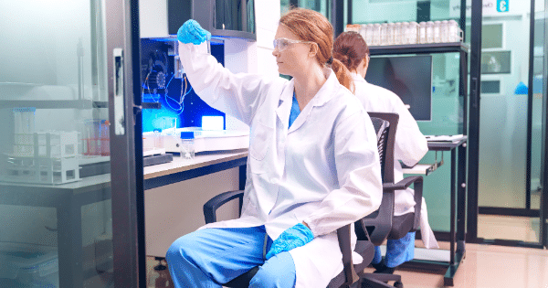 A female researcher wearing a lab coat, safety glasses, and blue gloves holds up a sample tube for inspection while seated at a workbench in a bright laboratory.