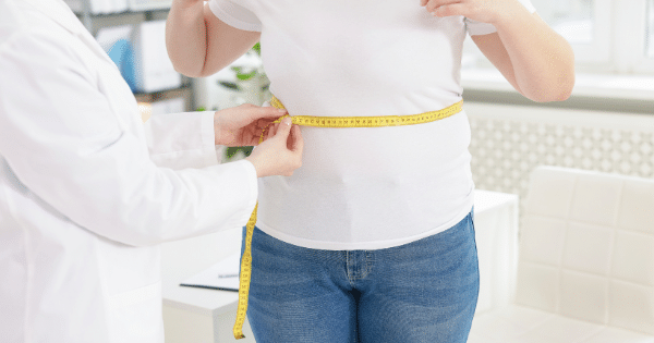 A medical professional in a white coat using a yellow measuring tape to measure the waist circumference of a person wearing a white t-shirt and blue jeans in a clinical setting.