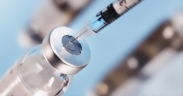 A close-up shot of a syringe needle inserting into the rubber stopper of a clear glass medication vial to draw out a blue-tinted liquid.