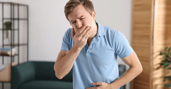 A man in a light blue polo shirt standing indoors, covering his mouth with one hand and clutching his stomach with the other, wearing an expression of discomfort or nausea.