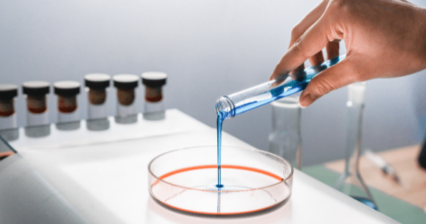 A gloved hand pouring a bright blue liquid from a test tube into a glass petri dish containing a reddish liquid on a lab workbench surrounded by other sample tubes.