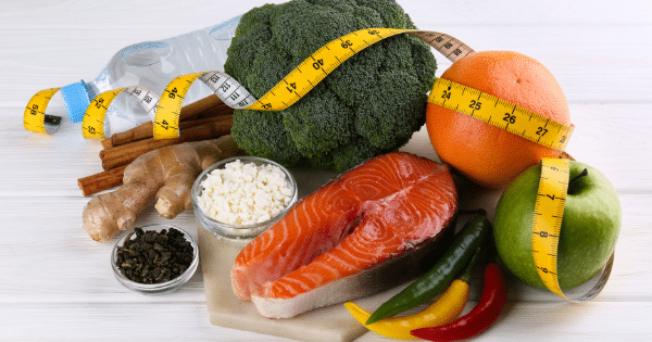 A healthy food arrangement on a white wooden surface featuring a salmon fillet, broccoli head, green apple, grapefruit, chili peppers, ginger root, cottage cheese, green tea, cinnamon sticks, a water bottle, and a yellow measuring tape wrapped around the items.