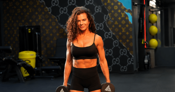 A fit, muscular woman with dark curly hair in a gym setting wearing a black sports bra and shorts, holding a dumbbell in each hand and looking directly into the camera.
