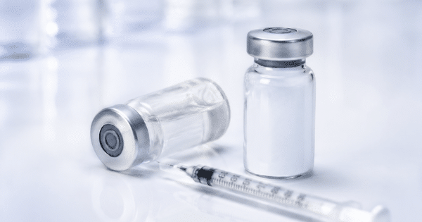A medical syringe laying next to two glass vials on a reflective white surface.