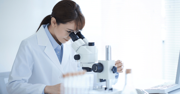 Female scientist in a lab coat looking through a microscope at a desk with test tubes.