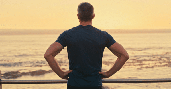 Rear view of a man with hands on his hips looking out over the ocean at sunset.