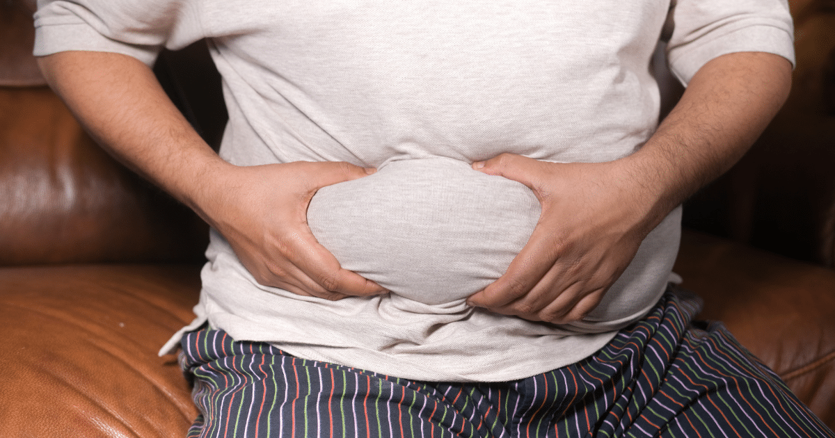 Close-up of a person sitting on a brown leather couch holding and squeezing a roll of excess belly fat through a light grey shirt.