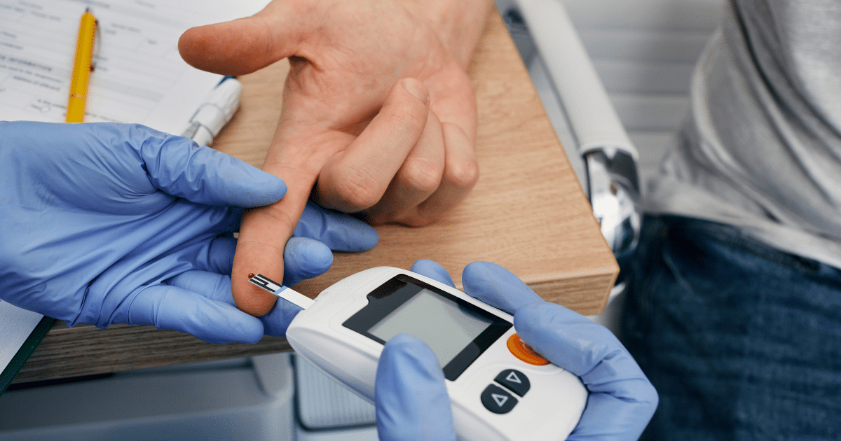 A gloved medical worker uses a blood glucose meter to test a small drop of blood on a patient's finger.