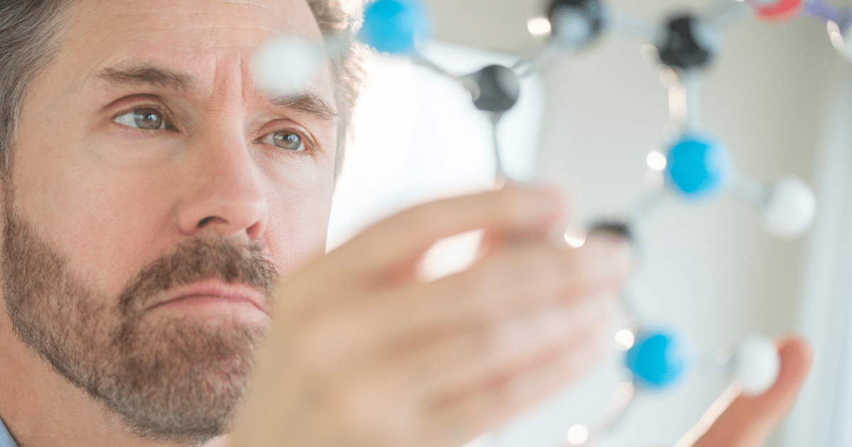 A middle-aged man with a beard and light brown hair looks thoughtful while examining a molecular structure model held in front of him.